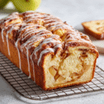 Amish Apple Fritter Bread 7 Freshly baked Amish Apple Fritter Bread loaf on a cooling rack, sliced slightly to reveal apple chunks and cinnamon swirl, drizzled with glaze.